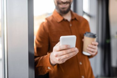 Ofiste tek kullanımlık kahve içerken şık gömlekli bir iş adamının cep telefonuyla mesajlaşmasının kısmi görüntüsü bulanık arka plan.
