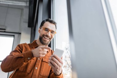 low angle view of excited entrepreneur with beard and eyeglasses, wearing stylish shirt, pointing with finger at mobile phone during video call in modern office  clipart