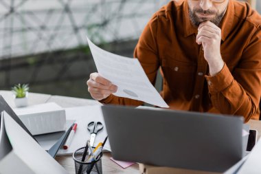 cropped view of thoughtful and busy businessman holding document near laptop, folders, scissors, marker and pen holder with pens while working at workplace in office clipart