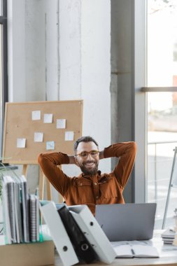 successful and joyful businessman in shirt and eyeglasses sitting with hands behind head in front of laptop, folders, notebooks and corkboard with sticky notes on background clipart