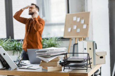 Meşgul ve yorgun iş adamı akıllı telefondan konuşurken mantar panonun yanında notlar ve not defterleri, kitaplar, karton kutular ve dizüstü bilgisayarlı çalışma masasıyla sorunu çözüyor.