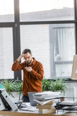 worried businessman in eyeglasses looking at mobile phone and holding hand near head next to work desk with laptop, notebooks, folders, books, paper cup and decorative plants in modern office clipart