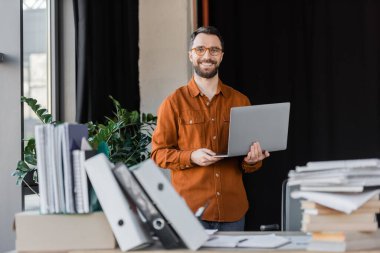 happy successful businessman in stylish eyeglasses and shirt standing with laptop and looking at camera near pile of books, notebooks and folders on blurred foreground in office clipart