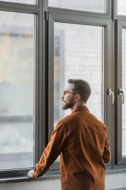brunette and bearded businessman in stylish eyeglasses and fashionable shirt, confident and successful, standing at large window in office and looking away clipart