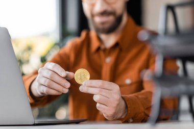 KYIV, UKRAINE - 18 Ekim 2022: memnun ve başarılı girişimcinin iş yerindeki çalışma masasının yanında altın bitcoin tutarken görüntüsü bulanık
