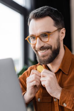 KYIV, UKRAINE - 18 Ekim 2022: memnun ve sakallı iş adamı şık gözlüklü, altın bitcoin tutan ve ofisteki bulanık önplanda dizüstü bilgisayara bakarken gülümseyen
