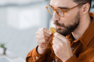 KYIV, UKRAINE - 18 Ekim 2022: modaya uygun gözlüklü, şık gömlekli, sakallı ve başarılı iş adamı çağdaş ofisin bulanık arka planında altın bitcoin tutuyor ve öpüyor.