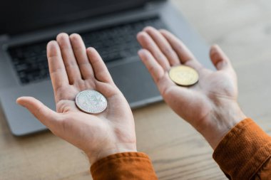 KYIV, UKRAINE - 18 Ekim 2022: Ofisteki bulanık dizüstü bilgisayarın yanındaki avuçlarında altın ve gümüş bitmadeni paralar tutan başarılı iş adamının kısmi görüntüsü