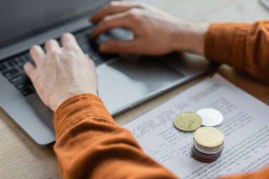 KYIV, UKRAINE - 18 Ekim 2022: Ofiste masasında altın ve gümüş bitcoin bulunan belgelerin yanındaki bulanık dizüstü bilgisayarda yazan başarılı iş adamının kısmi görüntüsü