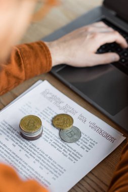 KYIV, UKRAINE - 18 Ekim 2022: Modern ofiste dizüstü bilgisayarda çalışan işadamının yanında kağıt belgeler üzerinde altın ve gümüş bitcoin