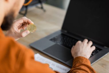 KYIV, UKRAINE - OCTOBER 18, 2022: partial view of blurred successful entrepreneur holding golden bitcoin and working on laptop with blank screen in office clipart