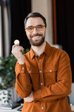 KYIV, UKRAINE - OCTOBER 18, 2022: charismatic and bearded businessman in trendy eyeglasses and shirt holding golden bitcoin and smiling at camera in modern office on blurred background clipart