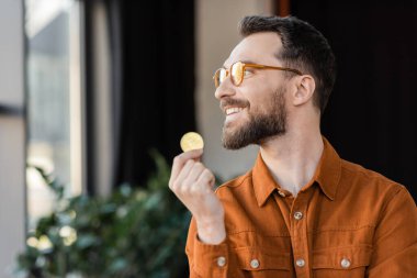 KYIV, UKRAINE - OCTOBER 18, 2022: joyful and accomplished businessman in stylish eyeglasses and shirt holding golden bitcoin, smiling and looking away in modern office on blurred background clipart