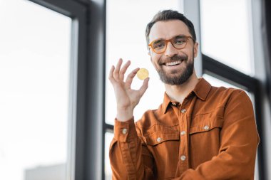 KYIV, UKRAINE - OCTOBER 18, 2022: bearded businessman in fashionable shirt and eyeglasses holding golden bitcoin and smiling at camera near blurred windows in modern office on blurred background clipart