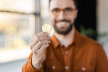 KYIV, UKRAINE - OCTOBER 18, 2022: cheerful, successful and bearded businessman in eyeglasses and shirt holding golden bitcoin and looking at camera in office clipart