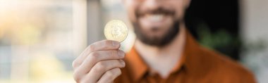 KYIV, UKRAINE - OCTOBER 18, 2022: selective focus of golden bitcoin in hand of cropped bearded businessman smiling on blurred background in office, banner clipart