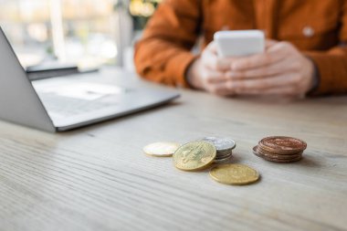 KYIV, UKRAINE - OCTOBER 18, 2022: selective focus of golden and silver bitcoins on desk near cropped businessman using mobile phone near laptop on blurred background in office clipart