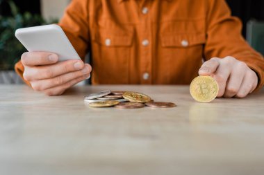 KYIV, UKRAINE - OCTOBER 18, 2022: partial view of entrepreneur in stylish shirt holding mobile phone while sitting near silver and golden bitcoins on work desk in office clipart