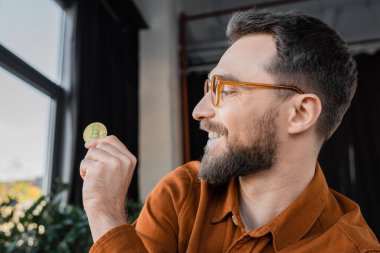 KYIV, UKRAINE - 18 Ekim 2022: esmer sakallı iş adamı, memnun ve başarılı, gözlük takıyor, bulanık arka plandaki çağdaş ofiste altın bitcoin 'e bakıyor