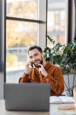Gülümseyen iş adamı sakala dokunuyor ve defterin yanında cep telefonuyla konuşuyor ve çağdaş ofiste dekoratif bitkilerin yanında bulanık dizüstü bilgisayar kullanıyor.