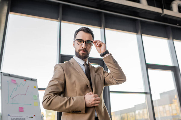 confident manager in stylish formal wear adjusting eyeglasses and looking at camera near flip chart with business planning and analytics on background 