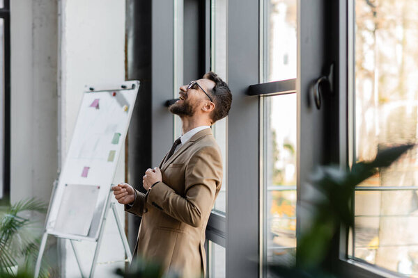 side view of overjoyed businessman in beige stylish blazer and eyeglasses leaning on windows and laughing while standing near flip chart with sticky notes in office