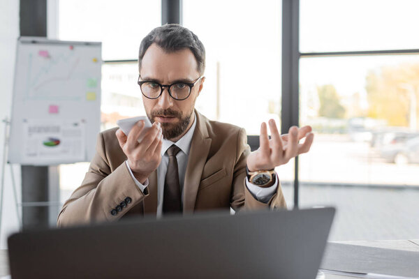 серьезный бизнесмен с брюнетками и бородой, в стильном бежевом пиджаке, галстуке и очках, смотрит на размытый ноутбук и отправляет голосовое сообщение на мобильный телефон