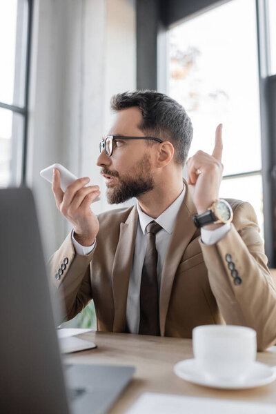 амбициозный бизнесмен в модном пиджаке, галстуке и стильных очках отправляет голосовое сообщение на смартфон и показывает жест внимания рядом с чашкой кофе и ноутбуком на размытом переднем плане