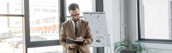 good looking and bearded corporate manager in eyeglasses and trendy business attire writing in notebook near flip chart with graphs while doing market research in modern office, banner