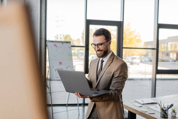 довольный предприниматель в стильной деловой одежде, стоящий с ноутбуком во время видеозвонка рядом с флип-чартом и рабочим столом с ноутбуком, документами и канцелярскими принадлежностями, размытый передний план