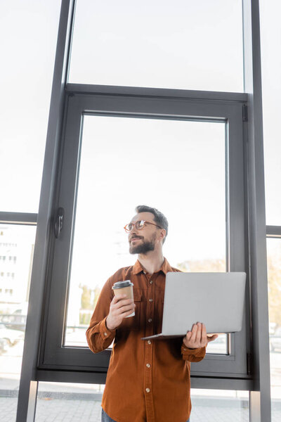 smiling and dreamy executive manager in stylish eyeglasses and shirt looking away while standing with laptop and coffee in paper cup near large office windows, corporate lifestyle 