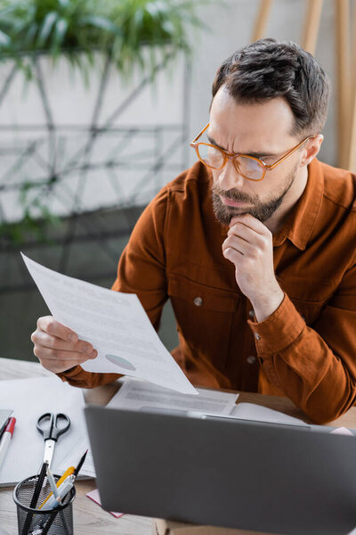 deep in thought bearded businessman in eyeglasses and shirt looking at document next to laptop, scissors and pen holder with pens on work desk in office