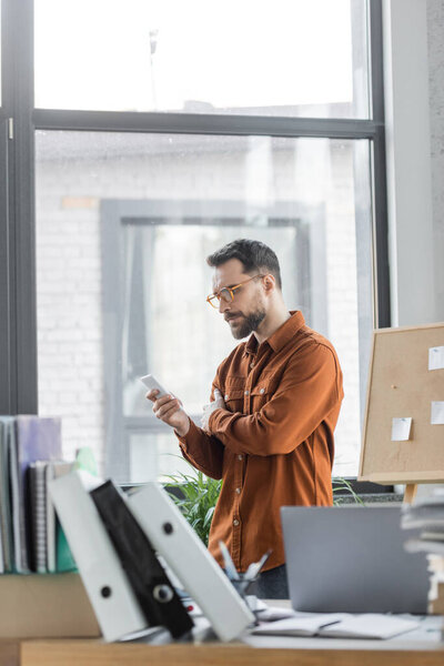 серьезный бородатый предприниматель в стильной рубашке и очках, глядя на смартфон рядом с рабочим столом с ноутбуком, много папок и ноутбуков на размытом переднем плане