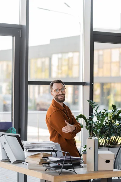 happy bearded businessman in eyeglasses and shirt smiling at camera while standing with crossed arms next to windows and workplace with pile of books, notebooks, folders, carton box and paper cup