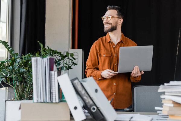 positive and successful entrepreneur in eyeglasses and shirt standing with laptop and looking away near blurred pile of books, notebooks and folders on work desk in office