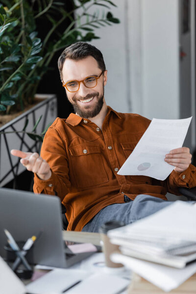 joyful bearded manager in stylish eyeglasses and shirt holding document and pointing at laptop during video conference near blurred documents on work desk in office
