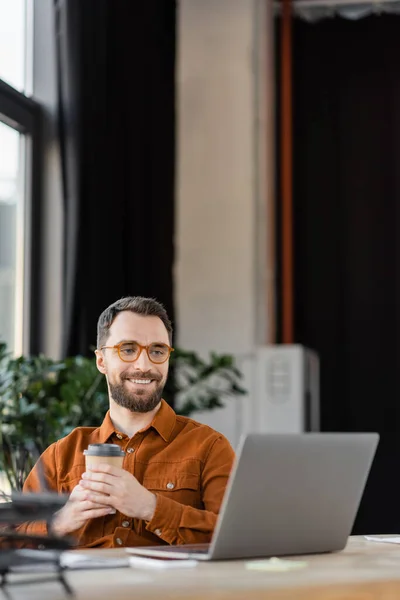 快乐的大胡子商人 戴着时髦的眼镜和衬衫 手里拿着咖啡 一边坐在办公室里 一边在模糊的背景下看着现代办公室的笔记本电脑 — 图库照片