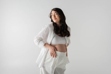 Beyaz şık gömlekli neşeli hamile kadın, bluz ve pantolonu göbeğe değiyor ve gri arka planda izole edilmiş, annelik moda konseptine bakıyor.