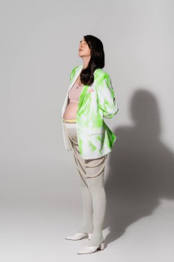 full length of pregnant woman with closed eyes standing in green and white jacket, crop top, beads belt and leggings on grey background, maternity style concept, expectation clipart