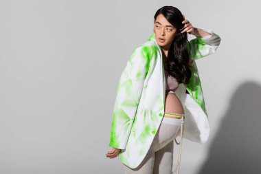 Müstakbel yeşil ve beyaz ceketli çekici anne tozluk ve boncuklu kemer dalgalı esmer saçlar ve gri arka plan, hamilelik moda konsepti, hamile kadın. 