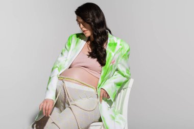 charming brunette mom-to-be in green and white blazer, crop top, leggings and beads belt sitting on chair on grey background, fashionable maternity concept, expectation clipart