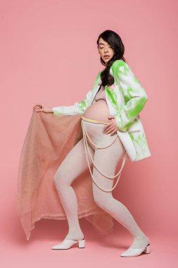 Uzun boylu, esmer, spor ceketli, açık tişörtlü, boncuk kemerli ve taytlı pembe arka planda bej şifon kumaşlı hamile bir kadın.