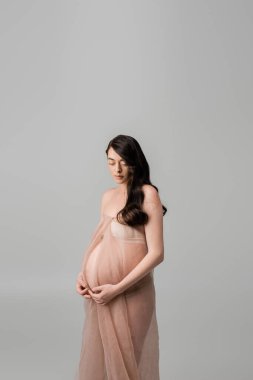 Gri arka planda izole edilmiş bej ve yumuşak şifon kumaşlar içinde duran kahverengi saçlı ve altın bilezikli romantik anne adayı, hamile kadın konsepti. 