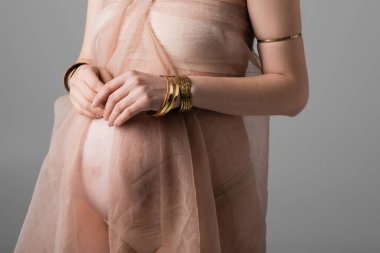 Yumuşak şifon kumaş ve altın aksesuarlı zarif ve şık hamile kadın manzarası gri arka planda izole edilmiş, annelik moda konsepti, şehvet, göbekli müstakbel anne.