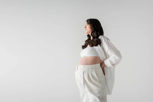 side view of smiling pregnant woman with wavy brunette hair, wearing white stylish maternity clothes, posing with hand behind back isolated on grey background, fashionable maternity concept