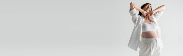 overjoyed pregnant model in white crop top, shirt and pants stretching arms with closed eyes isolated on grey background, maternity fashion concept, banner