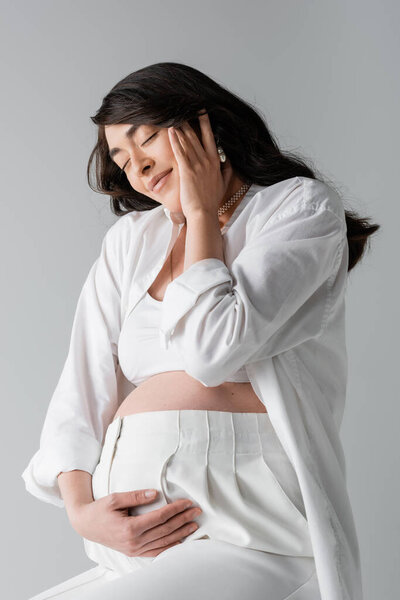 brunette pregnant woman in white crop top, shirt and pants touching belly and happy face with closed eyes isolated on grey background, maternity style concept