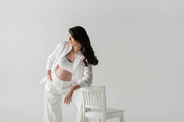 pleased pregnant woman in fashionable pants, crop top and shirt leaning on chair and looking away isolated on grey background, maternity fashion concept