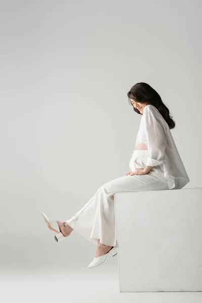 幸せなブルネットの母となるの側面図グレーの背景にスタジオでキューブに座っている白いシャツやパンツなどのファッショナブルな服 マタニティファッションの概念 — ストック写真