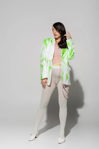 full length of future mother in leggings, beads belt, crop top and green and white blazer looking away while standing on grey background, trendy pregnancy concept